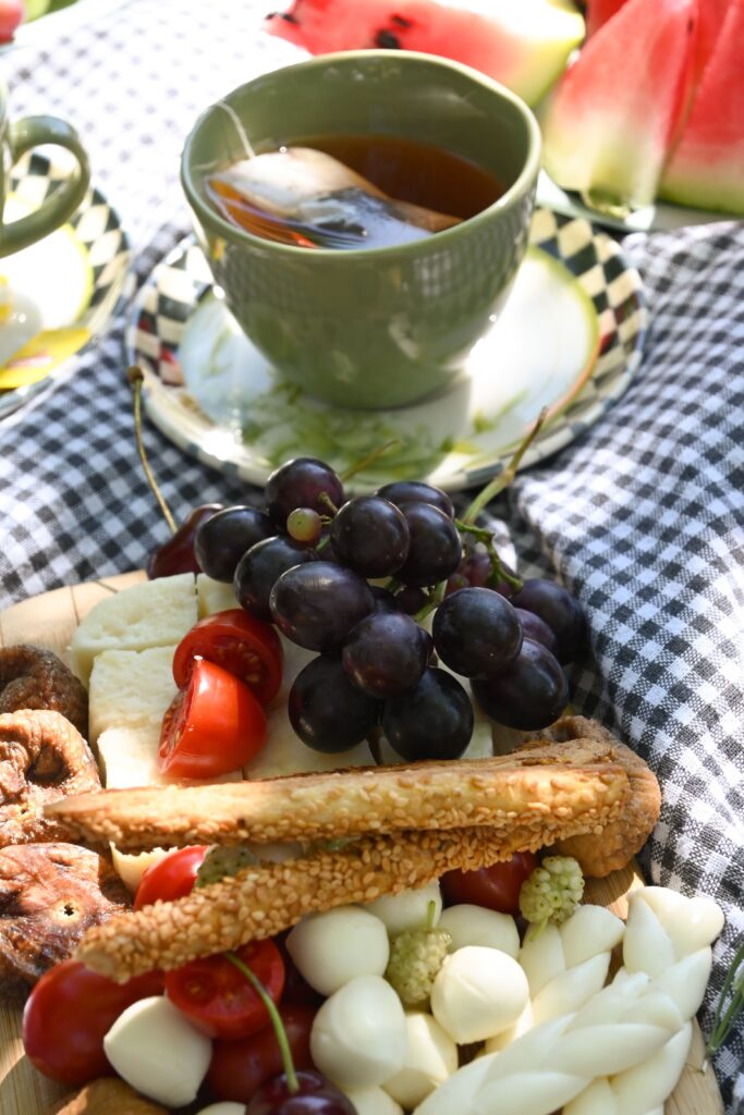 Das Foto zeigt leckes Essen auf einer karierten Picknickdecke. Zu sehen sind Brotstangen mit Sesam, Kirschtomaten, Mozzarella-Kugeln, getrocknete Feigen, dunkelrote Weintrauben, in Scheiben geschnittene Wassermelone und eine Tasse Tee.